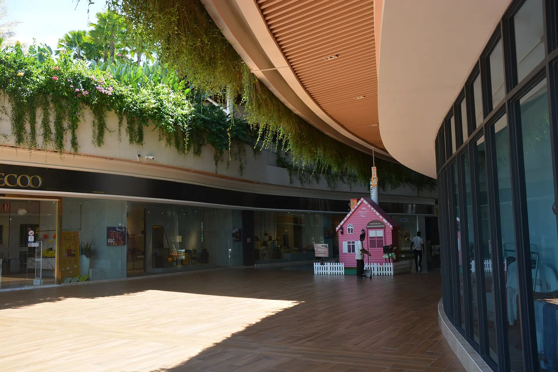 A shaded commercial street inside Forest City, Johor with people, greenery, and storefronts.