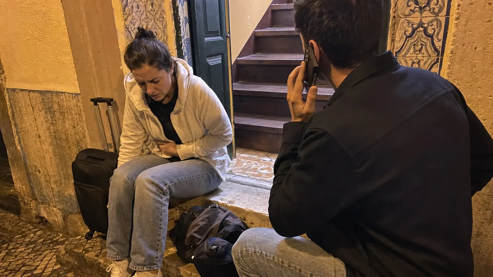 Candid phone photo of a traveler with abdominal pain sitting near luggage in a Lisbon guesthouse hallway.